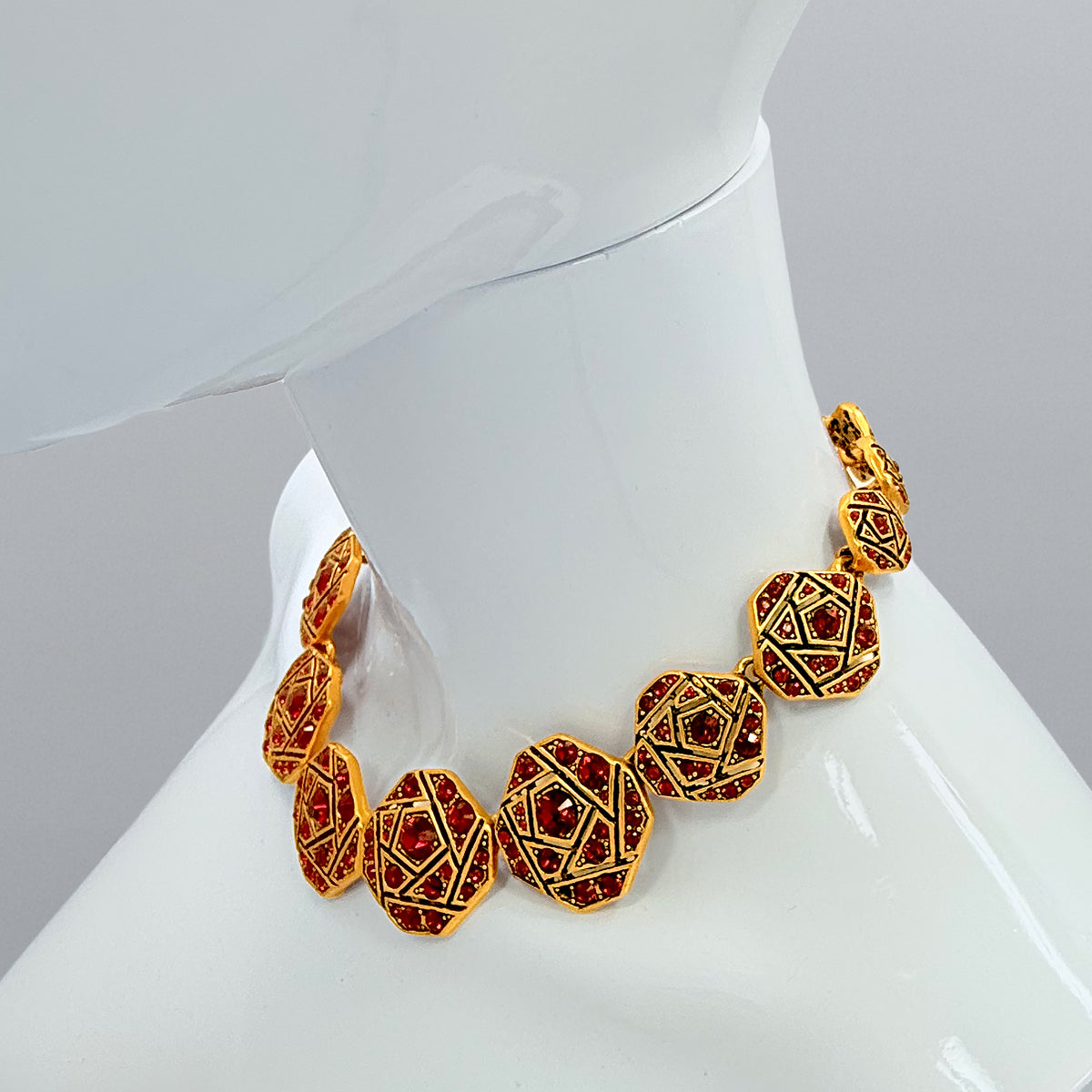 A close-up of Oscar de la Renta's geometric mosaic necklace featuring Red Siam Austrian crystals set in antique gold-plated frames, styled with a red dress for a bold statement look.