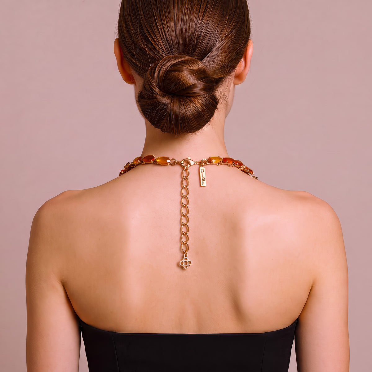 A woman shown from the back wearing a gold chain necklace with amber-colored beads and a small hanging tag, against a soft mauve background.