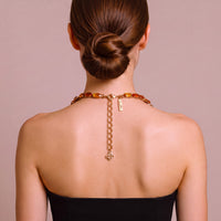 A woman shown from the back wearing a gold chain necklace with amber-colored beads and a small hanging tag, against a soft mauve background.